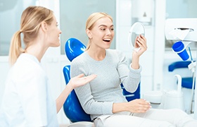 Woman with blond hair in blue dental chair smiling at her reflection