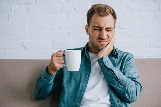 Squinting man holding white mug in one hand and touching sore jaw with the other
